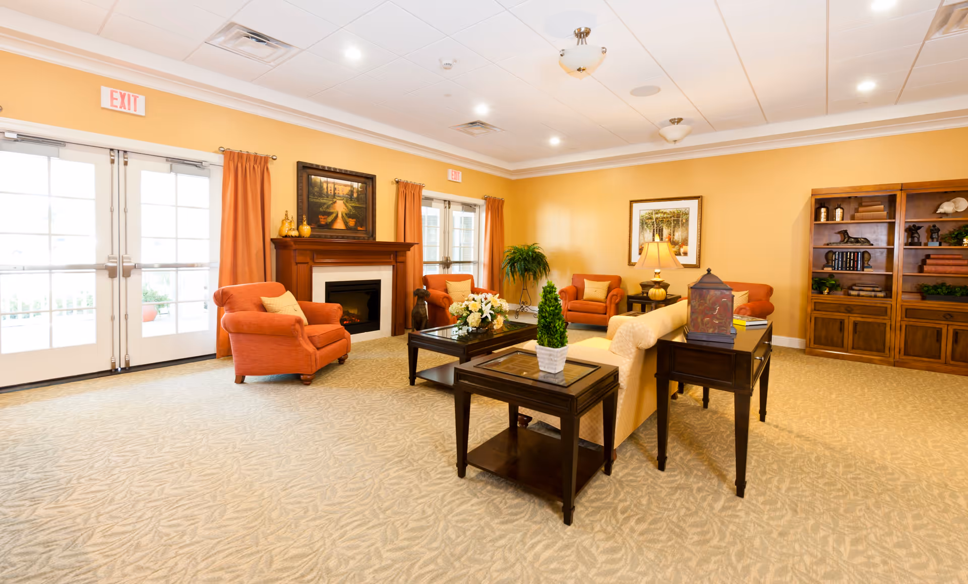 A bright and spacious living room area with beige carpet and yellow walls. The room features a fireplace with a painting above it, two orange armchairs, a beige sofa, wooden side tables with decorative items, a lamp, a large wooden bookshelf, and large windows with orange curtains letting in natural light.