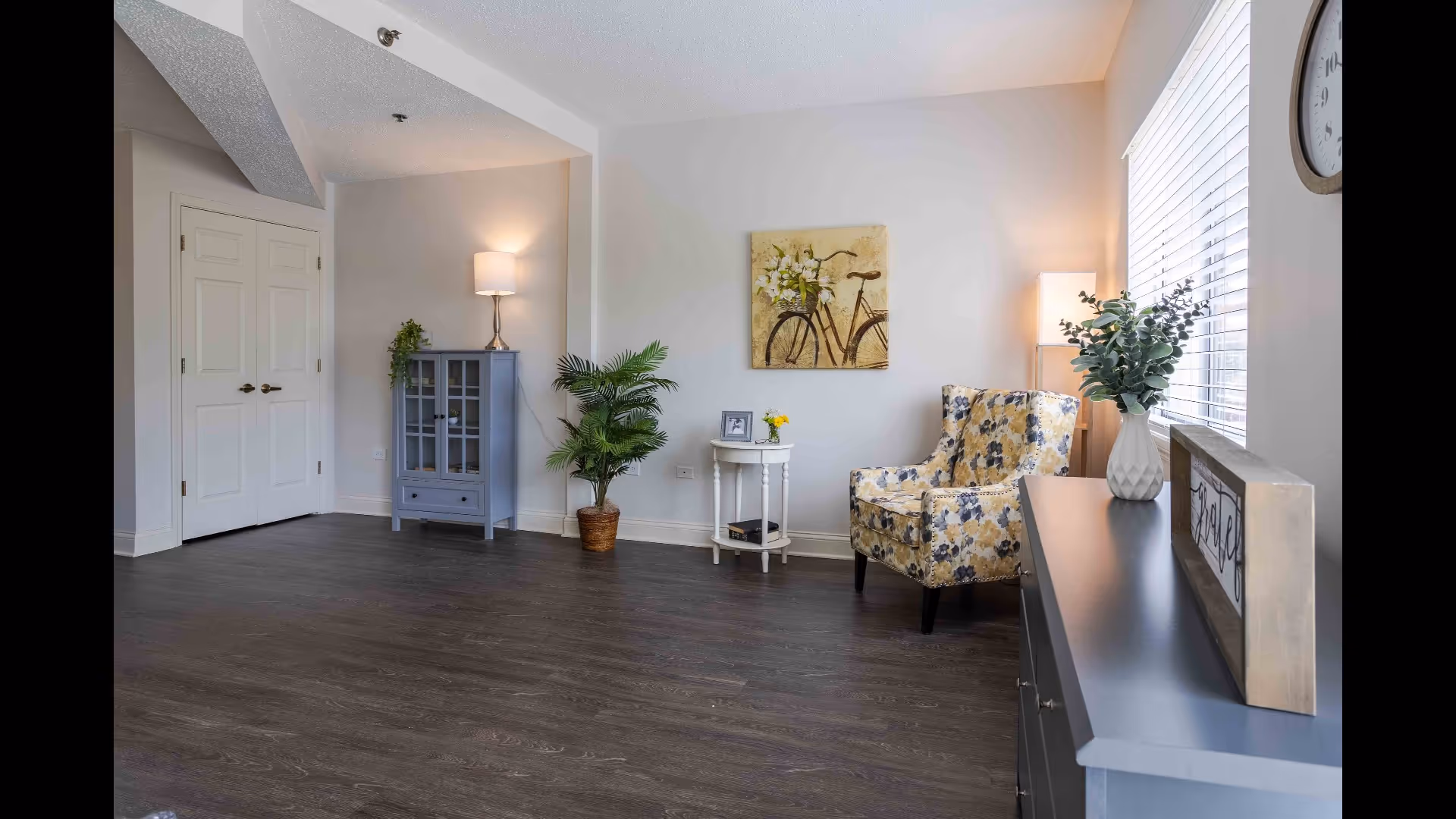A bright and cozy living room area with dark wood flooring, a floral patterned armchair, a small white side table with a photo frame and yellow flowers, a blue cabinet with a lamp on top, a potted plant, and a window with blinds letting in natural light. A framed picture of a bicycle with flowers hangs on the wall above the armchair. A gray console table with a vase of greenery and a decorative sign is positioned near the window.