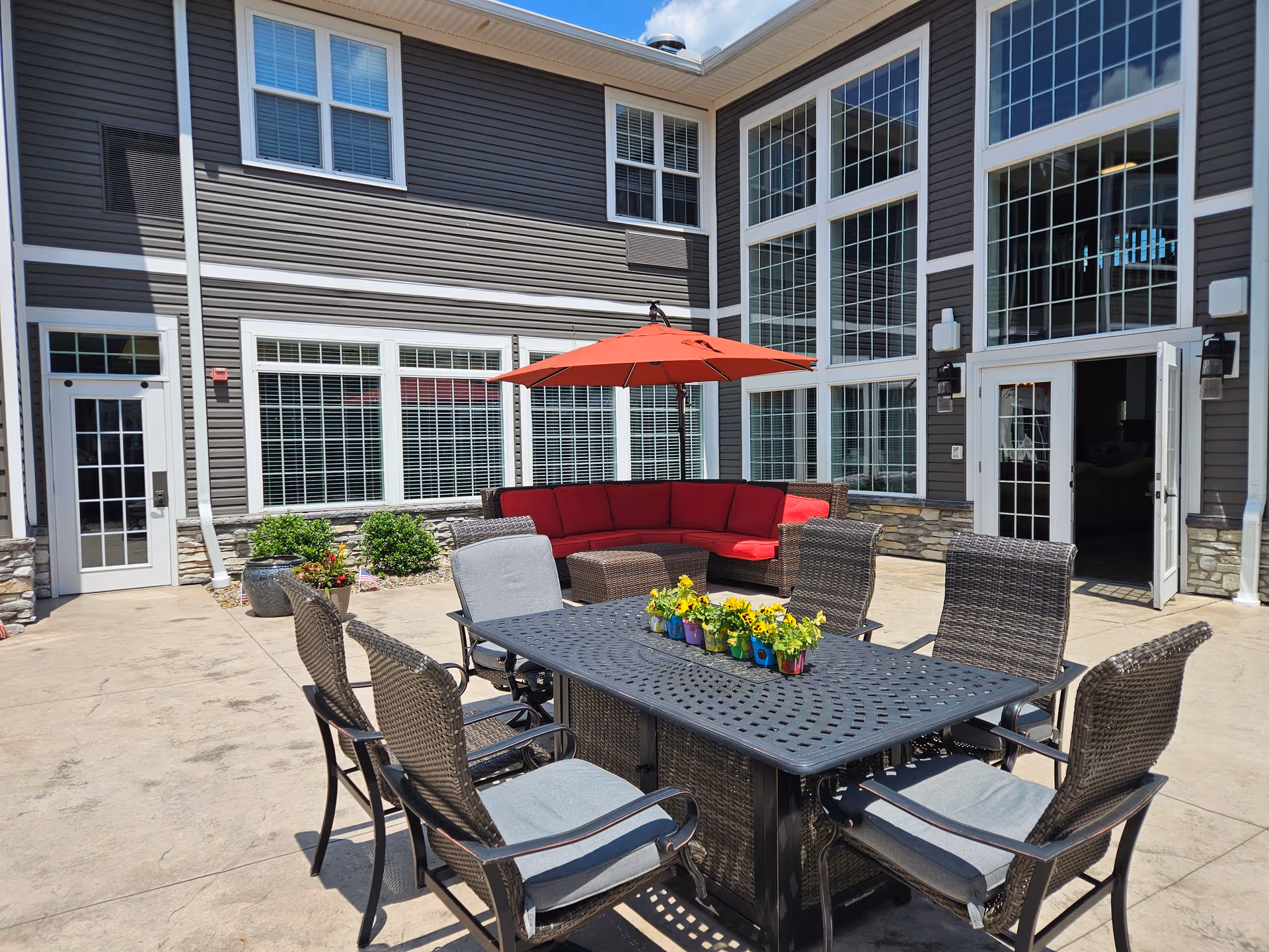 Outdoor patio courtyard with a dining table and wicker chairs, a red umbrella and sectional sofa in front of a multi-story building.