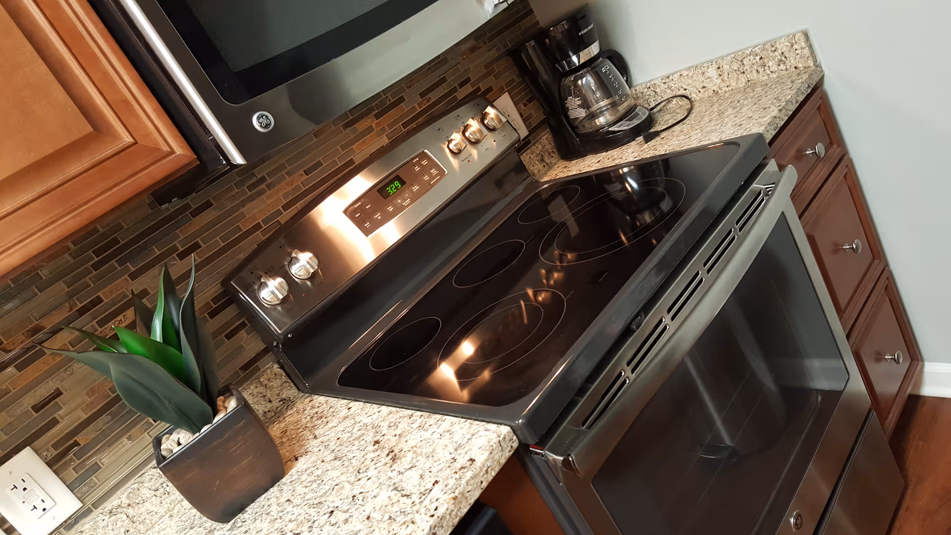 Modern kitchen countertop with a black electric stove and oven, a coffee maker, a small potted plant, and a tiled backsplash. The cabinetry is wooden with silver knobs.