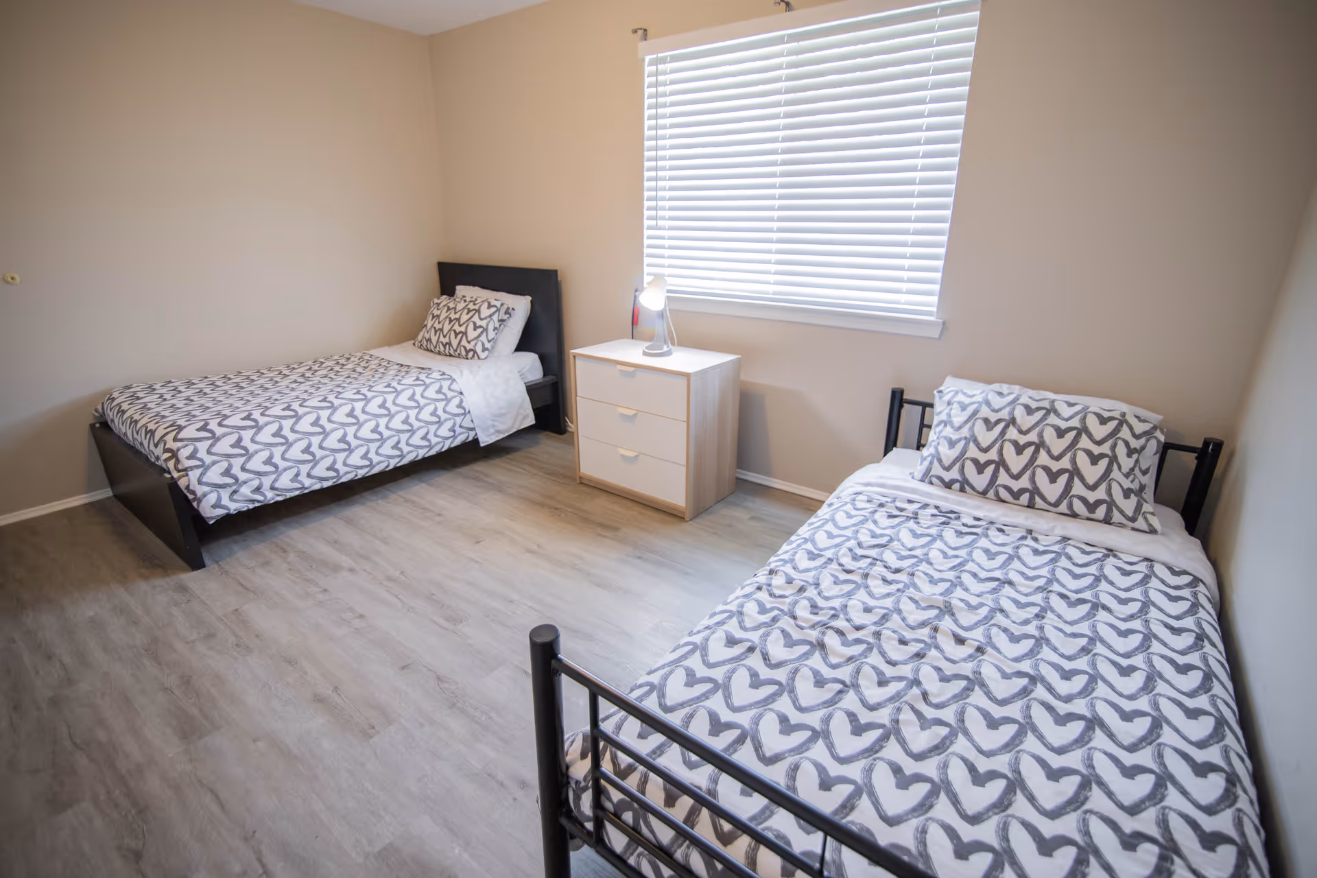 A simple bedroom with two single beds, each with white bedding featuring a gray heart pattern. Between the beds is a small white nightstand with three drawers and a small lamp on top. A window with white blinds is on the wall behind the nightstand. The room has light-colored walls and wood flooring.