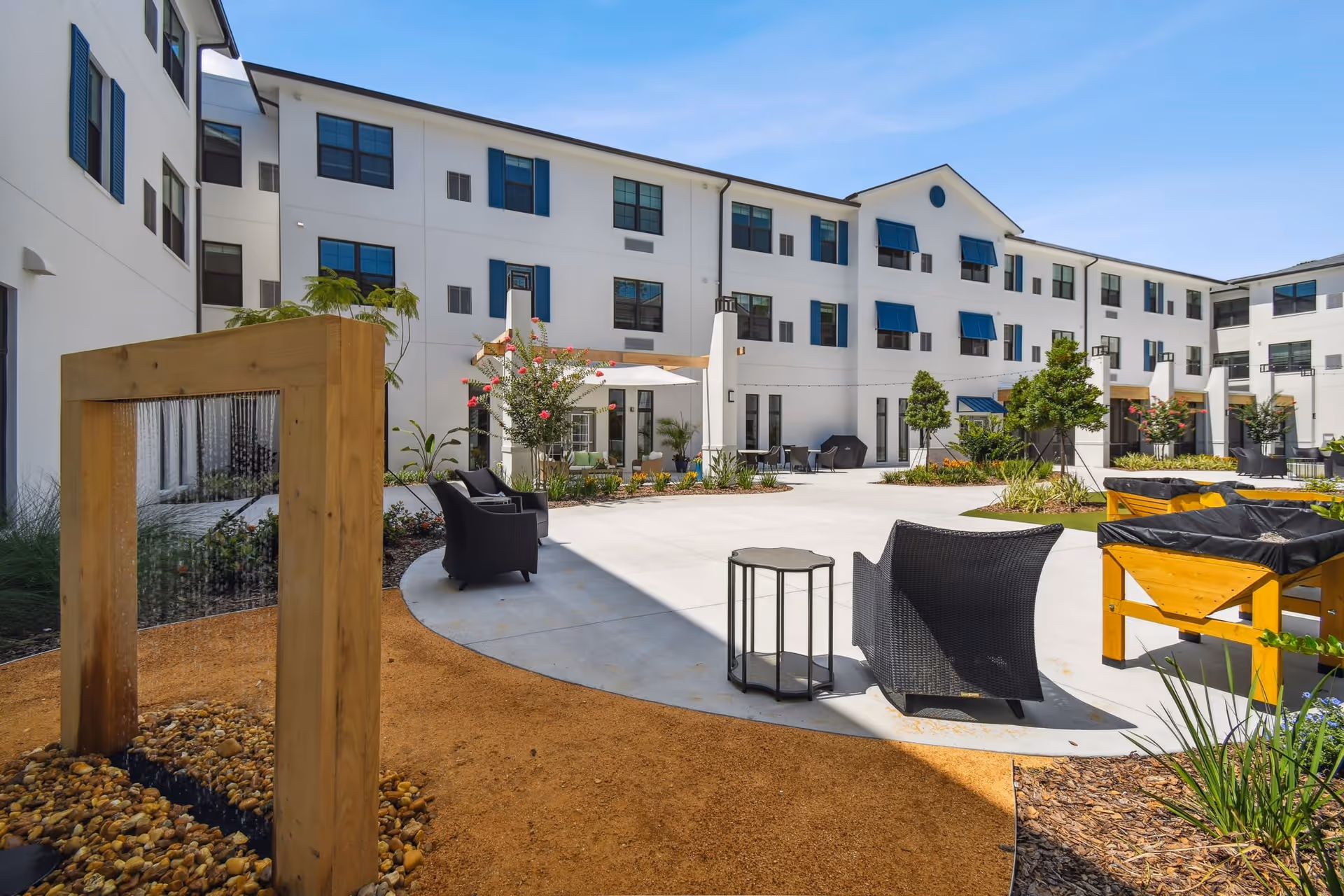 Outdoor courtyard area of a senior living facility with modern white buildings featuring blue window shutters and awnings. The courtyard includes black wicker chairs, small tables, raised garden beds, a wooden water feature, and landscaped plants under a clear blue sky.