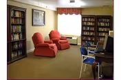 Cozy communal reading room with bookshelves, two red armchairs, a window with a valance, and a small desk with chairs.