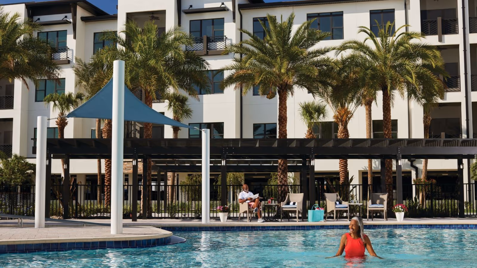 Outdoor swimming pool area at Siena Lakes Senior Living Community with a woman in a red swimsuit standing in the pool and a man sitting on a chair reading a newspaper. Palm trees and a multi-story building are visible in the background.