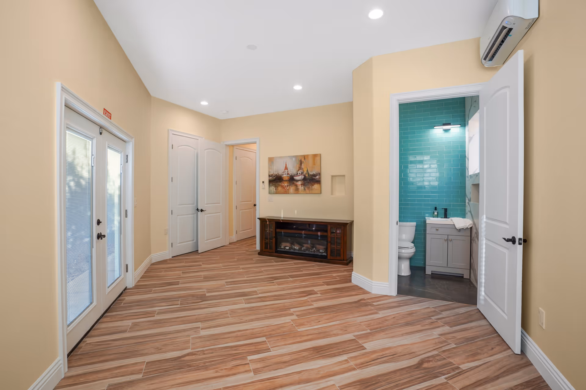 Interior view of a senior living facility room with light beige walls and wood-patterned tile flooring. The room features double glass doors on the left, two closed white doors in the center, and an open door on the right revealing a bathroom with turquoise tiled walls, a toilet, and a vanity with a sink. A painting hangs on the wall above a dark wooden electric fireplace.