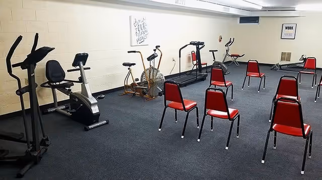 A fitness room with exercise equipment including stationary bikes, elliptical machines, and a rowing machine. Several red chairs are arranged in rows on a carpeted floor. The walls are white with motivational posters.