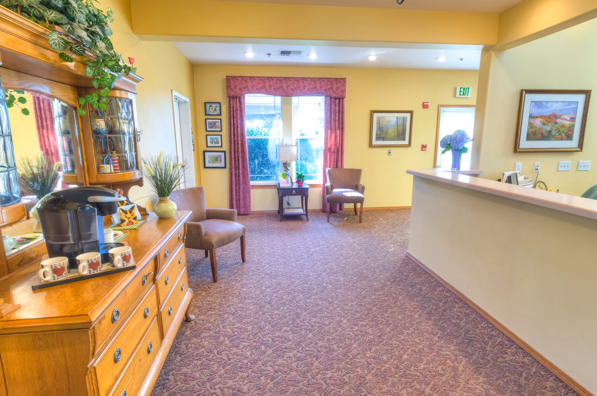 A bright and welcoming reception area with a wooden dresser holding a coffee maker and mugs on the left. Two upholstered chairs are positioned near a window with red curtains. The reception desk is on the right side with a vase of flowers and framed artwork on the walls. The carpet is patterned and the walls are painted yellow.