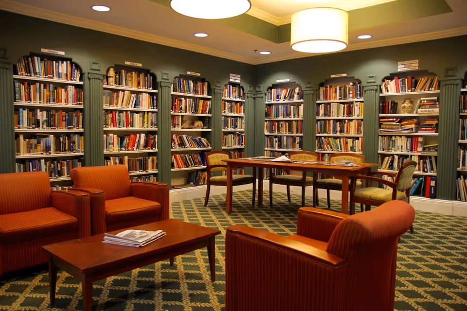 A cozy library room with green walls and carpet, featuring multiple bookshelves filled with books. There are several orange armchairs and a wooden coffee table with newspapers on it in the foreground. In the background, a wooden table with chairs is positioned in front of the bookshelves. The room is softly lit by ceiling lights and a large hanging lamp.