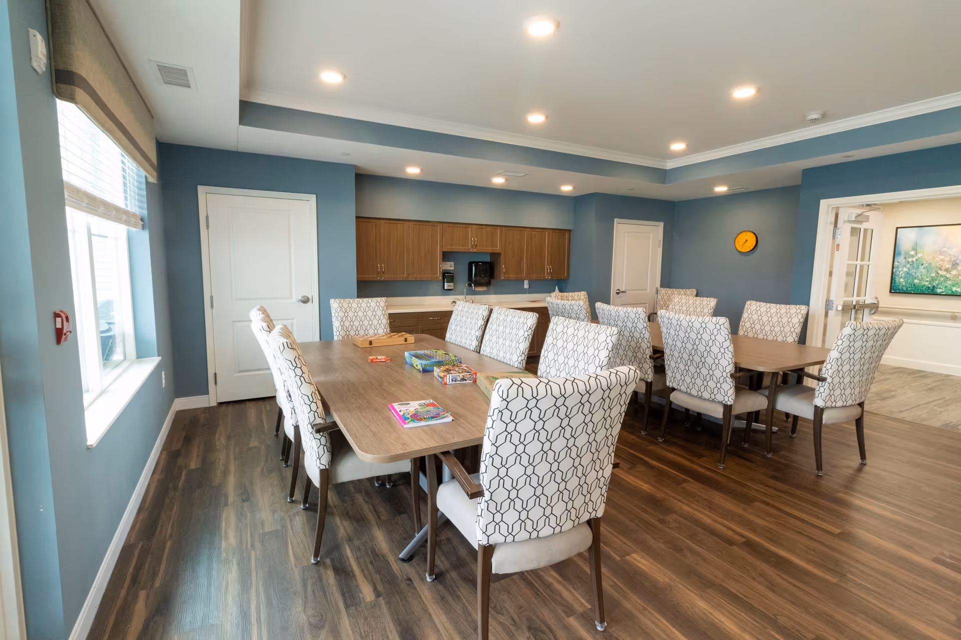 A well-lit room with blue walls and wood flooring featuring two rectangular tables surrounded by cushioned chairs with patterned upholstery. On the tables are various board games and activity books. The room has recessed ceiling lights, a clock on the wall, and cabinets with a countertop along one side. A window with blinds is on the left, and two closed white doors are visible.