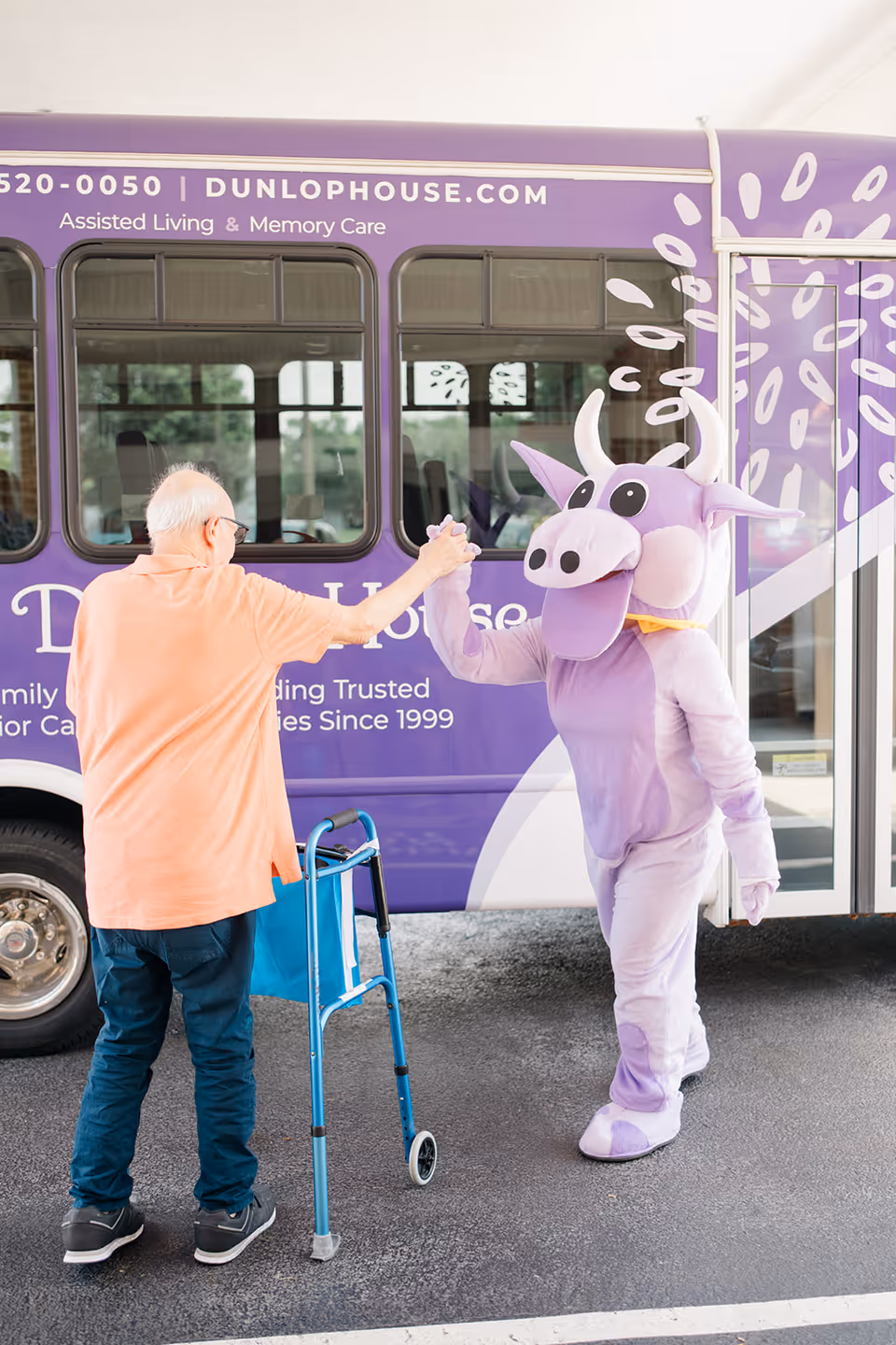 An elderly man with a walker high-fives a person in a purple cow mascot costume beside a purple Dunlop House shuttle bus.