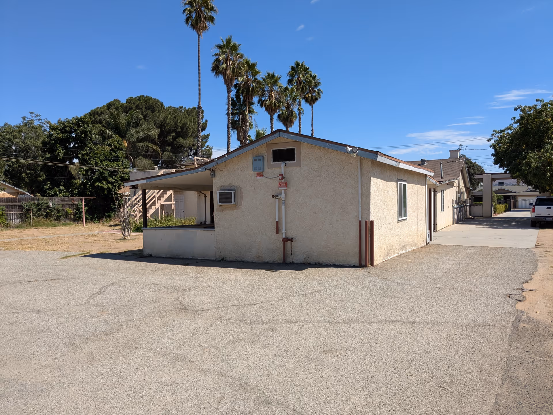 Single-story stucco building next to a paved lot with palm trees in the background under a clear blue sky.