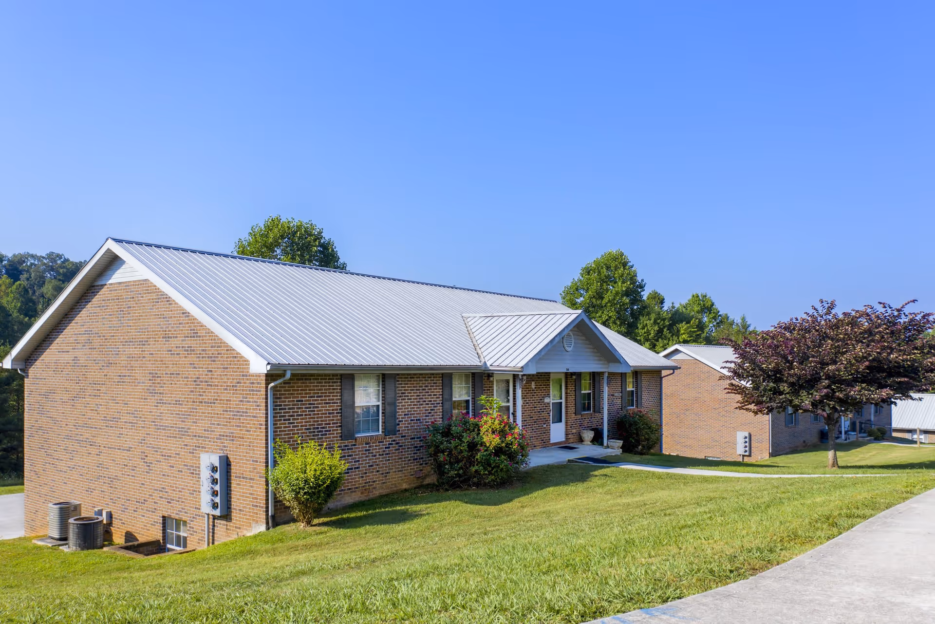 Exterior view of a single-story brick building with a metal roof at Small Town Senior Living at Deer Ridge. The building is surrounded by a well-maintained lawn, bushes, and a tree, with a clear blue sky overhead.