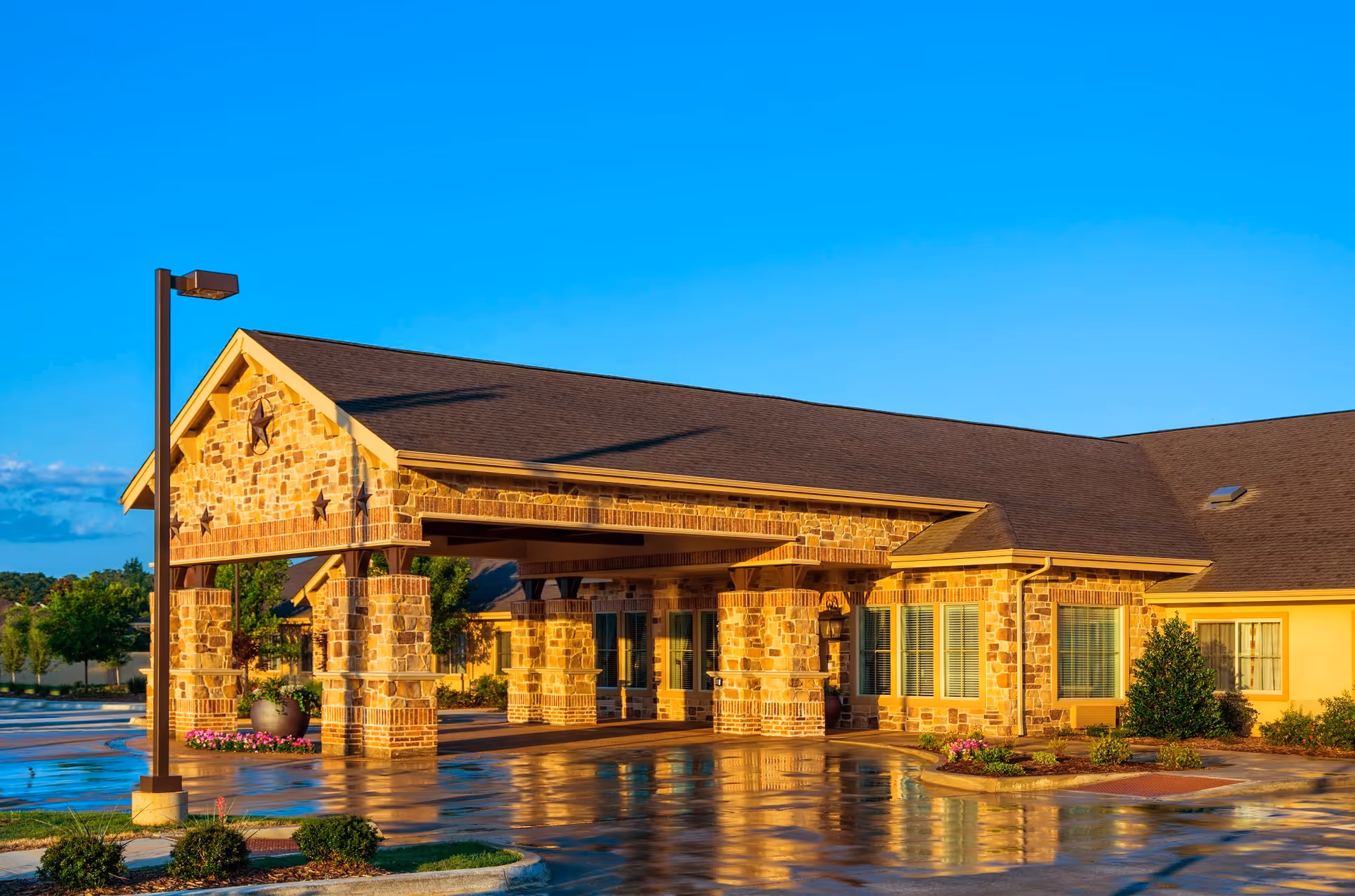 Exterior view of a single-story building with stone and brick facade, featuring a covered entrance supported by stone pillars. The area in front is wet, reflecting the building and the clear blue sky above.