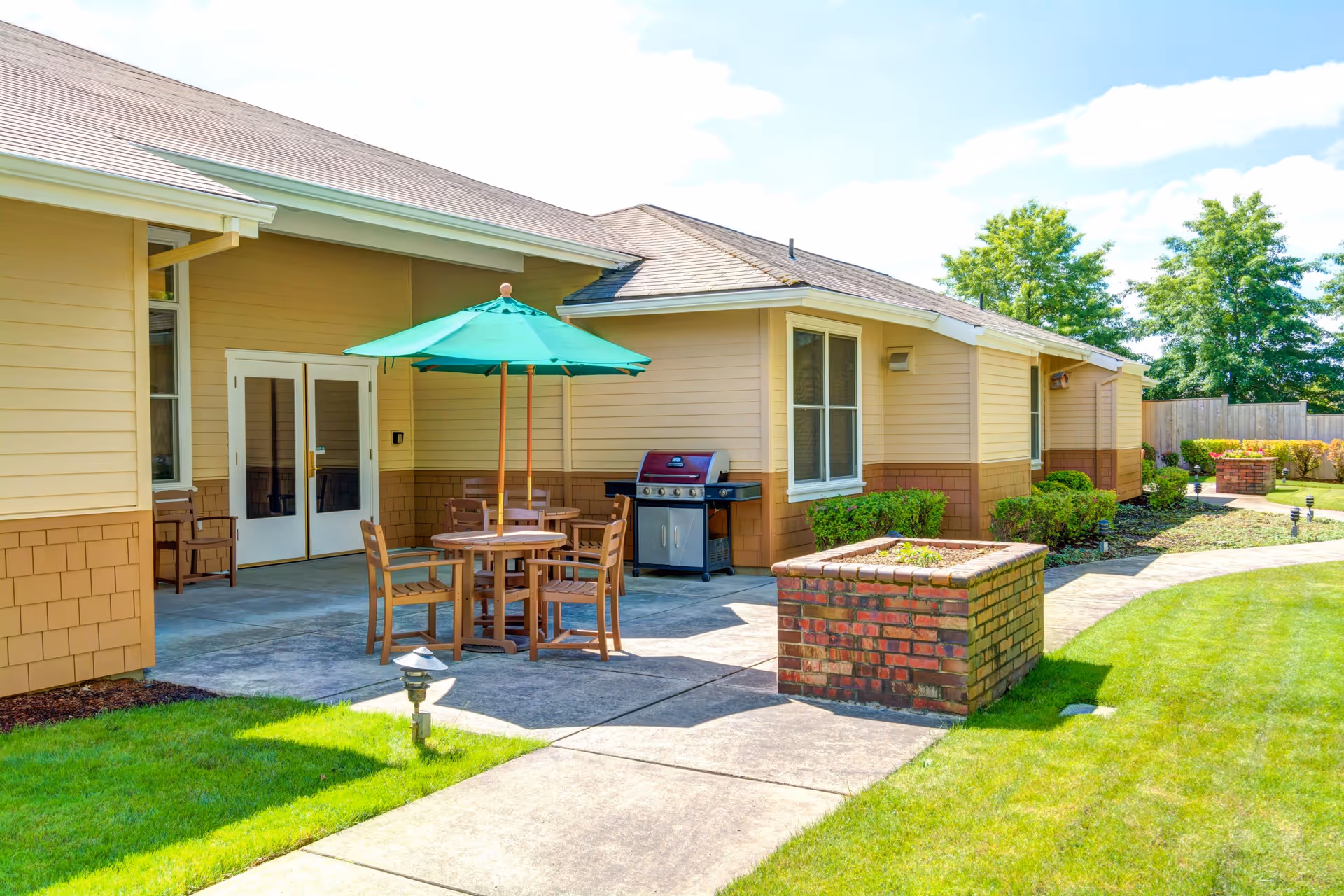 Outdoor patio area at Monterey Court Memory Care featuring a round wooden table with four chairs and a green umbrella. There is a barbecue grill against the building wall, surrounded by well-maintained grass, shrubs, and a brick planter. The building has beige siding with brown accents and white-framed windows and doors.