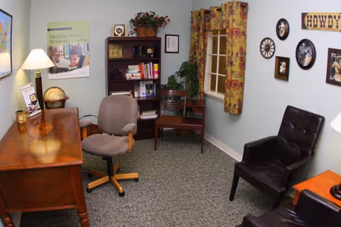 A cozy office or study room with a wooden desk, a swivel chair, two leather armchairs, a bookshelf filled with books and decorative items, a window with floral curtains, and wall decorations including framed pictures and a sign that says 'HOWDY'.