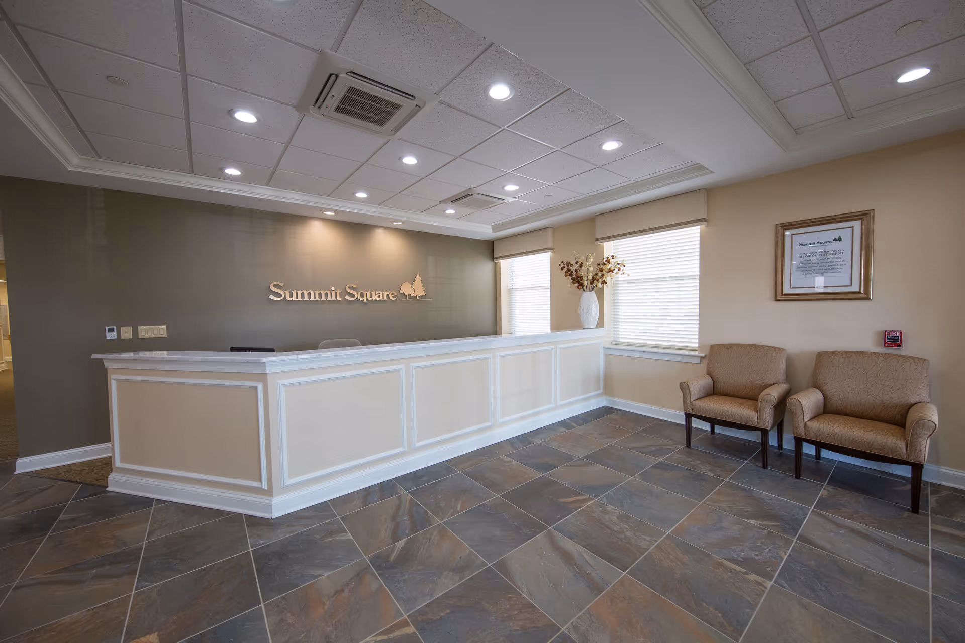 Reception area of Summit Square Retirement Community featuring a white reception desk, a wall with the facility's name and logo, two beige armchairs, a large vase with dried flowers, and windows with blinds allowing natural light into the room.