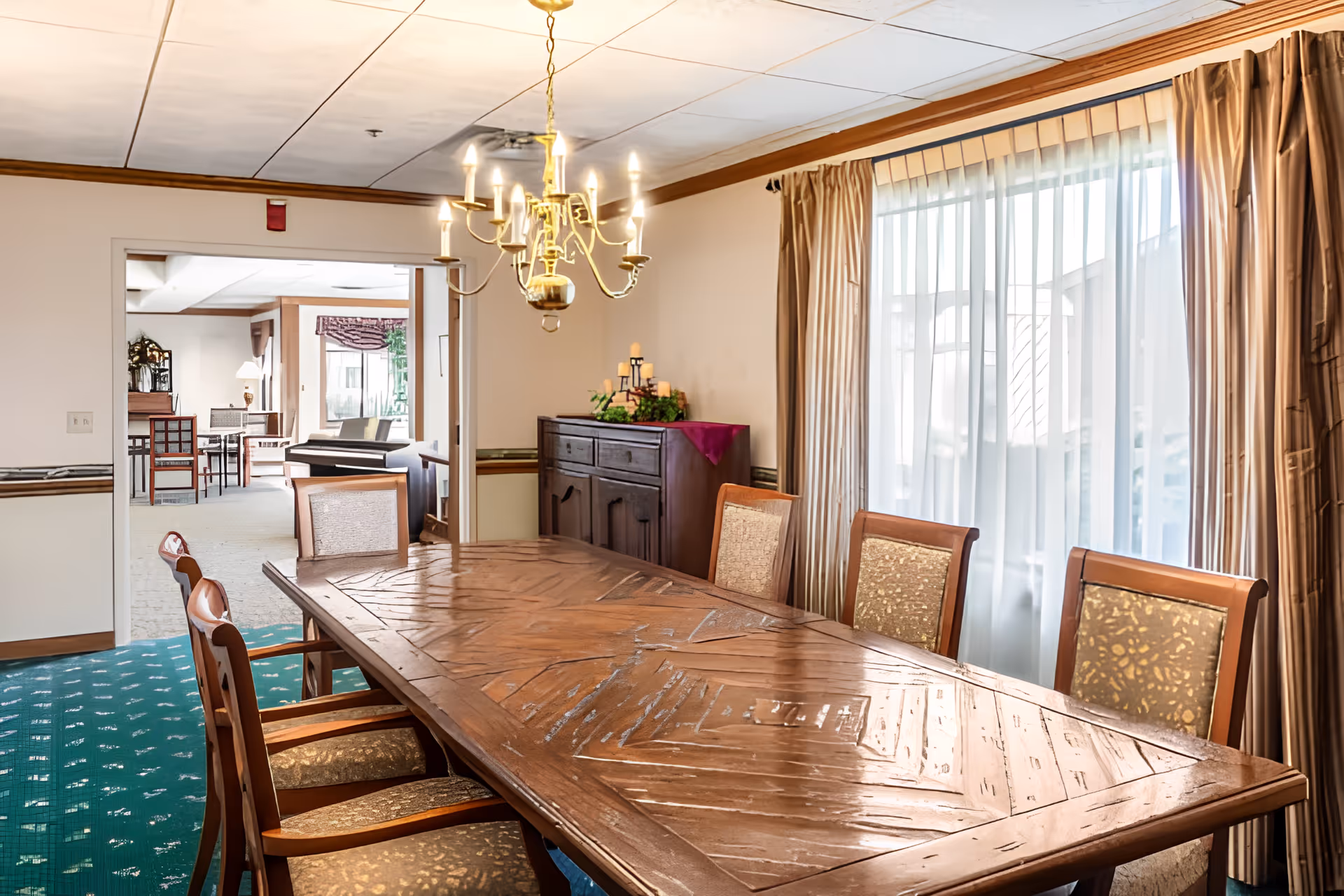 Dining room with a long wooden table surrounded by upholstered chairs, a chandelier, sideboard, and large curtained windows.