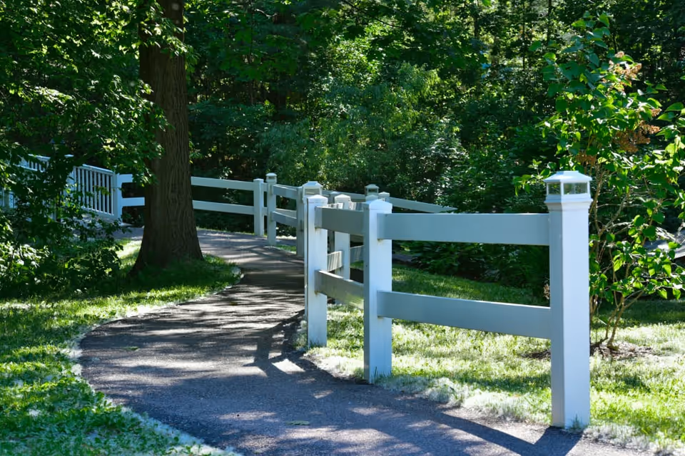 A winding paved pathway bordered by white wooden fences on both sides, surrounded by lush green trees and grass in a peaceful outdoor setting.