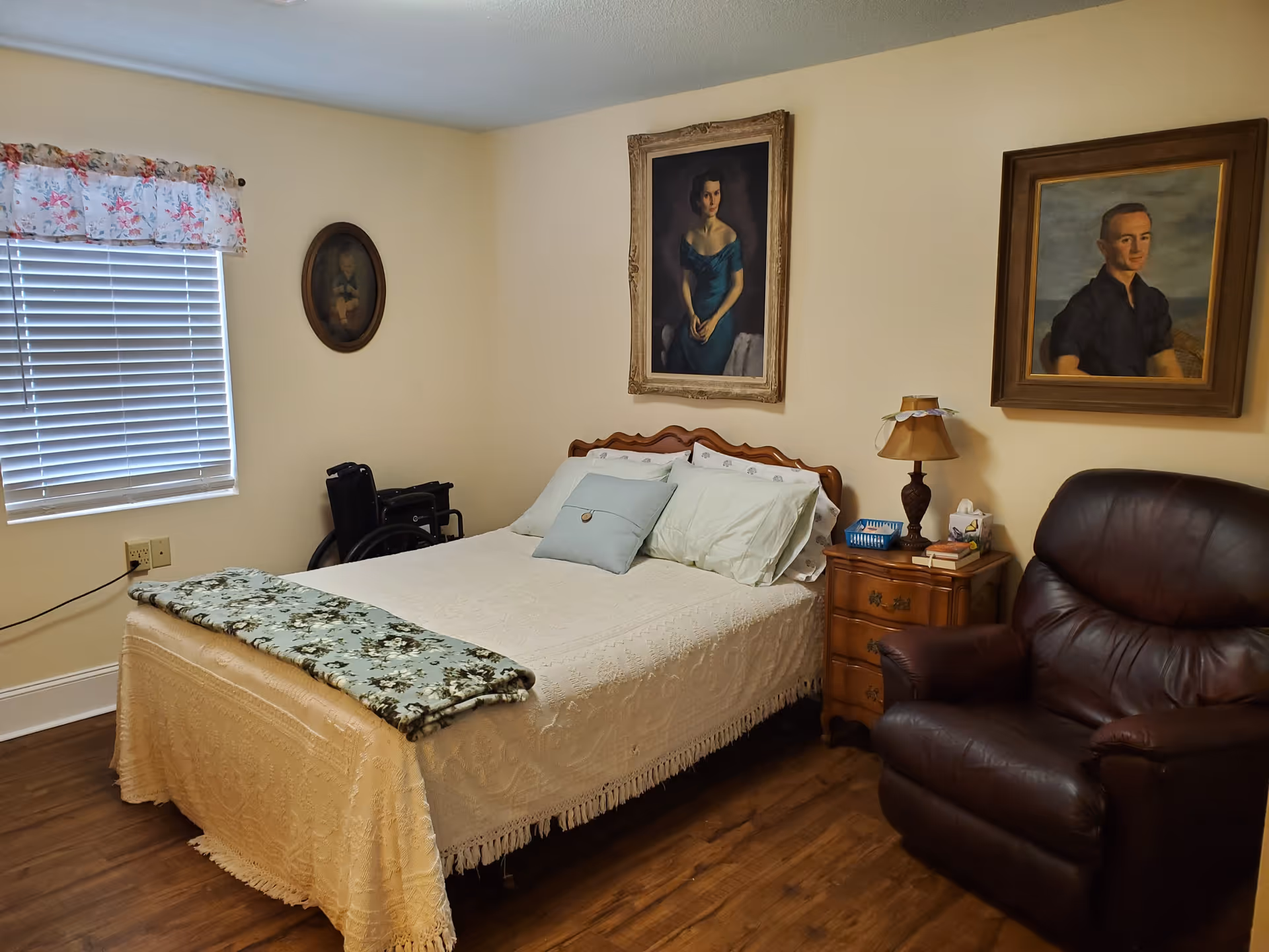 A neatly furnished bedroom with a made bed, bedside table and lamp, leather recliner, wheelchair, and framed portraits on the walls.