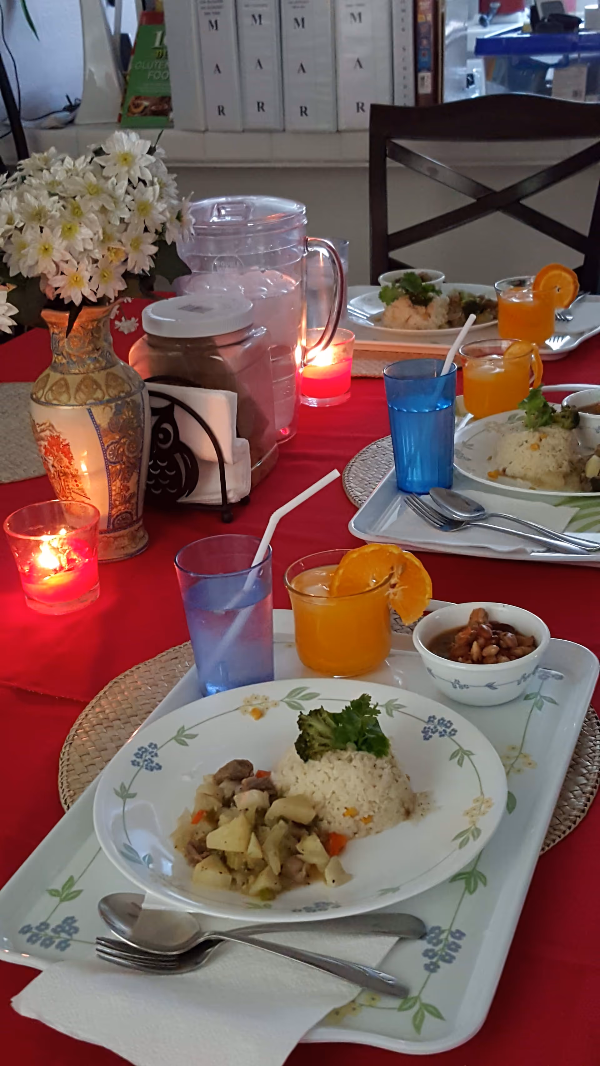 A dining table set with trays of food including rice, vegetables, and beans, accompanied by glasses of water and orange juice with a slice of orange. The table is covered with a red tablecloth and decorated with a vase of white flowers and lit candles. Chairs and shelves with binders are visible in the background.