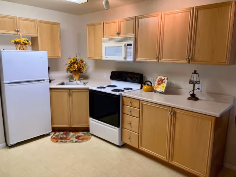 A kitchen with light wood cabinets, a white refrigerator, a white microwave above a white electric stove with black cooktop, a small sink with a floral arrangement, a yellow kettle on the stove, a decorative plate, and a small lamp on the countertop.