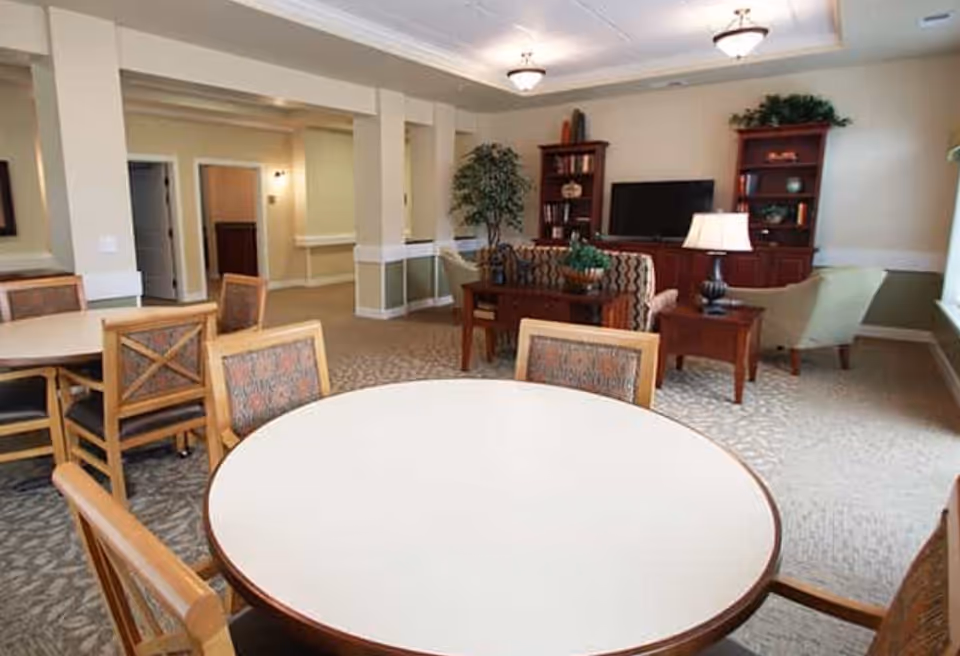 Communal dining and lounge area with round tables and chairs in the foreground and a TV with bookshelves and seating in the background.