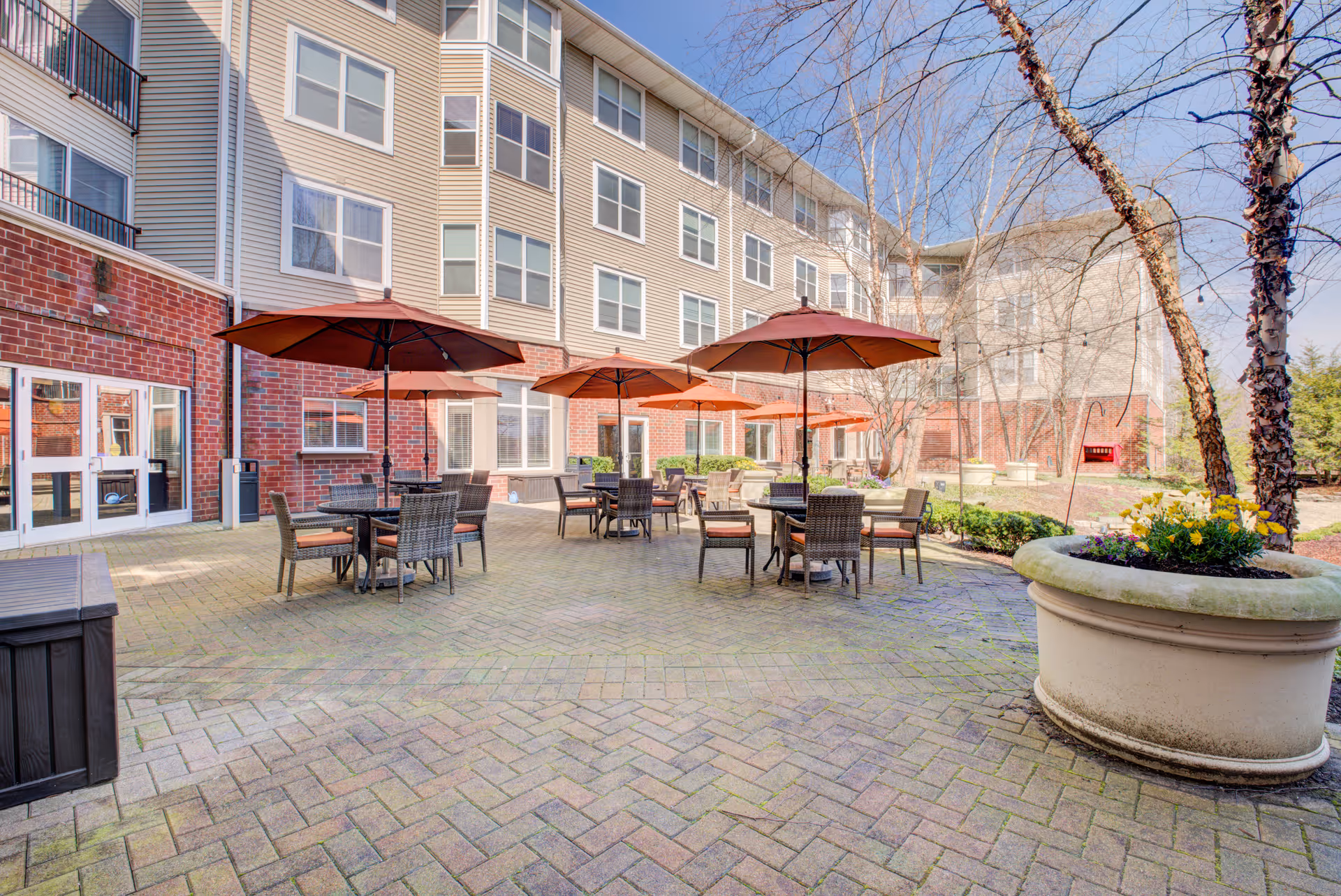 Outdoor patio area at Lake Barrington Woods with several tables and chairs under large orange umbrellas, surrounded by a multi-story building with brick and siding exterior. There are large planters with flowers and some leafless trees, under a clear blue sky.