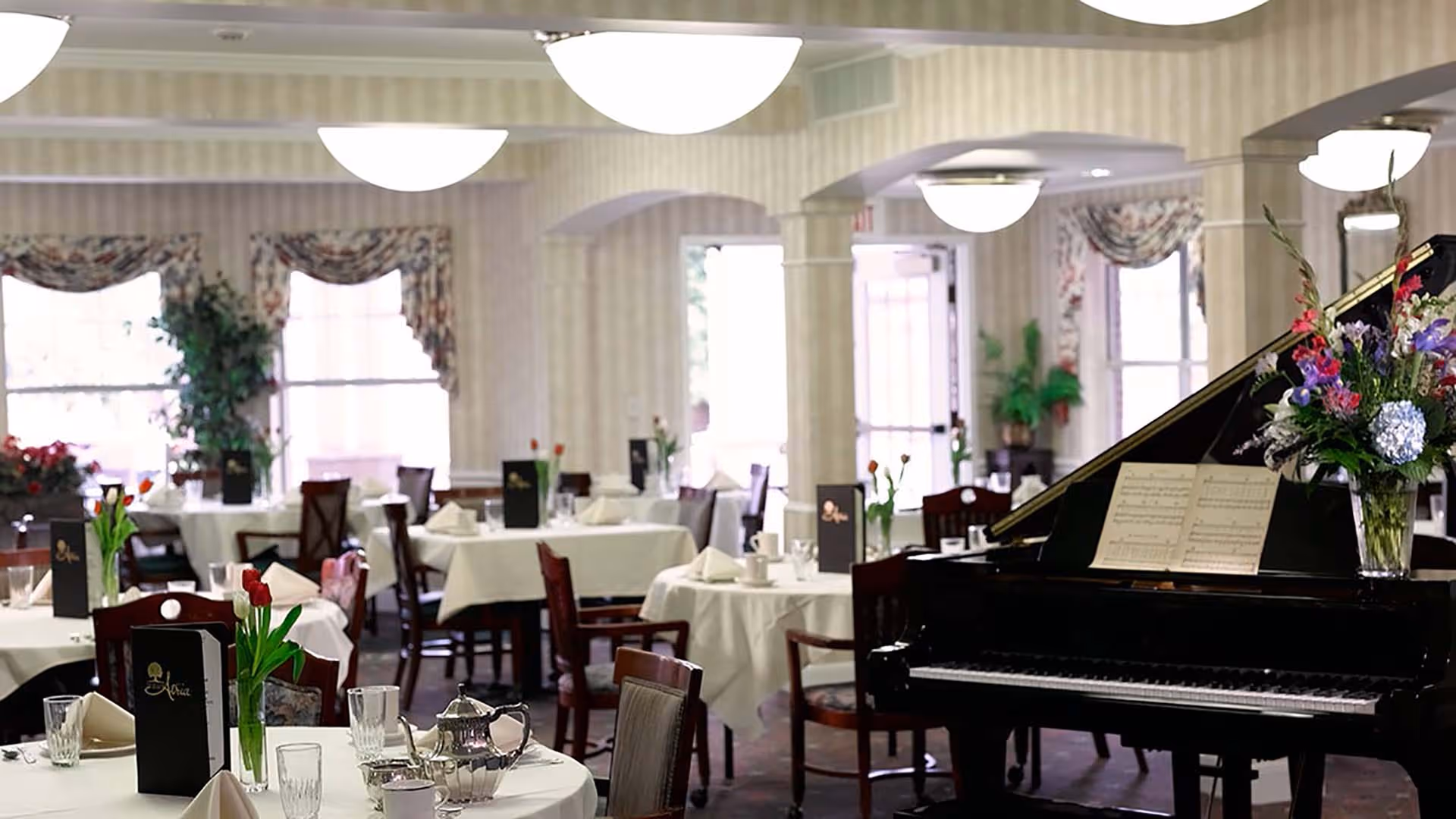 Elegant dining room with white-clothed tables set for service and a grand piano topped with sheet music and flowers.