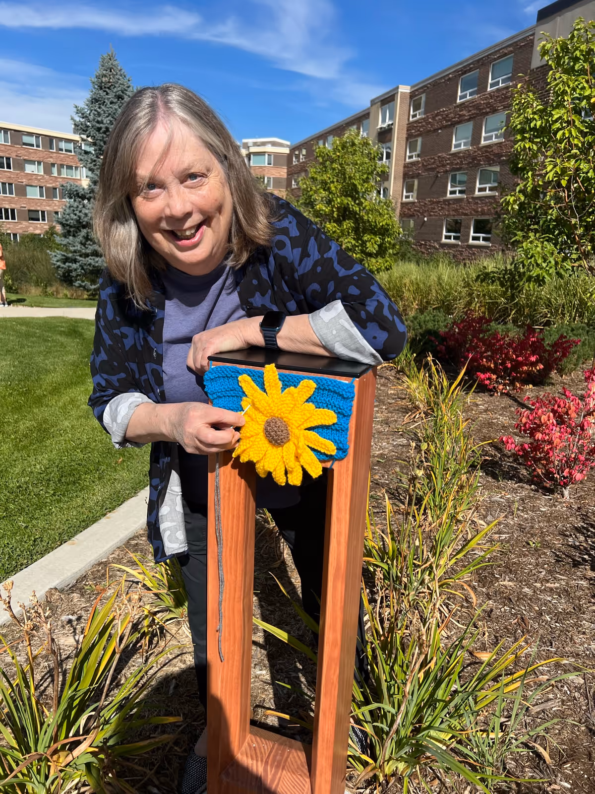 A smiling woman with shoulder-length gray hair stands outdoors in a garden area near a wooden post decorated with a knitted blue and yellow sunflower. Behind her are green bushes, a tree, and a multi-story brick building under a clear blue sky.
