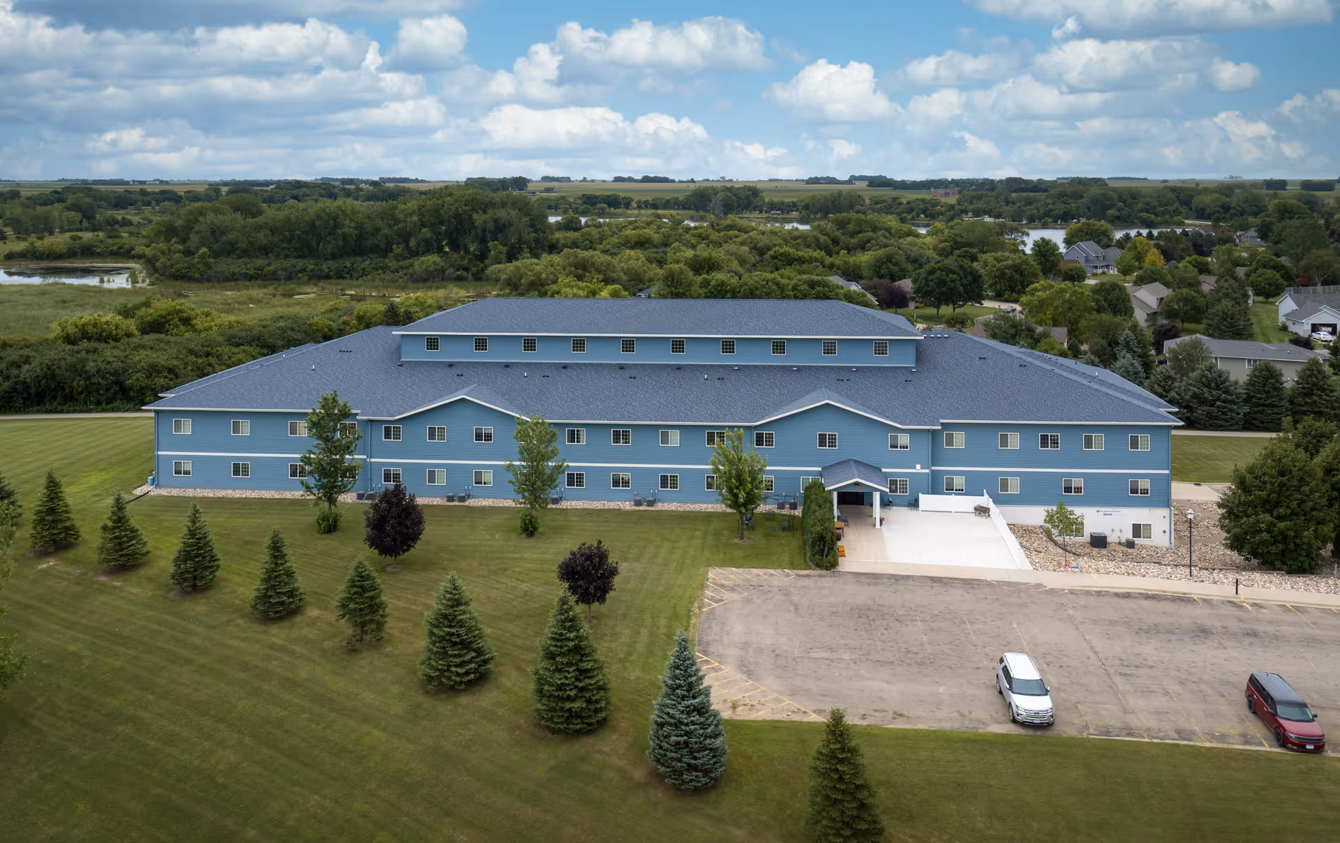 Aerial view of a large blue three-story building with a dark roof, surrounded by green lawns and trees. There is a parking area with a few cars in front of the building. The background shows a rural landscape with more trees, water bodies, and scattered houses under a partly cloudy sky.