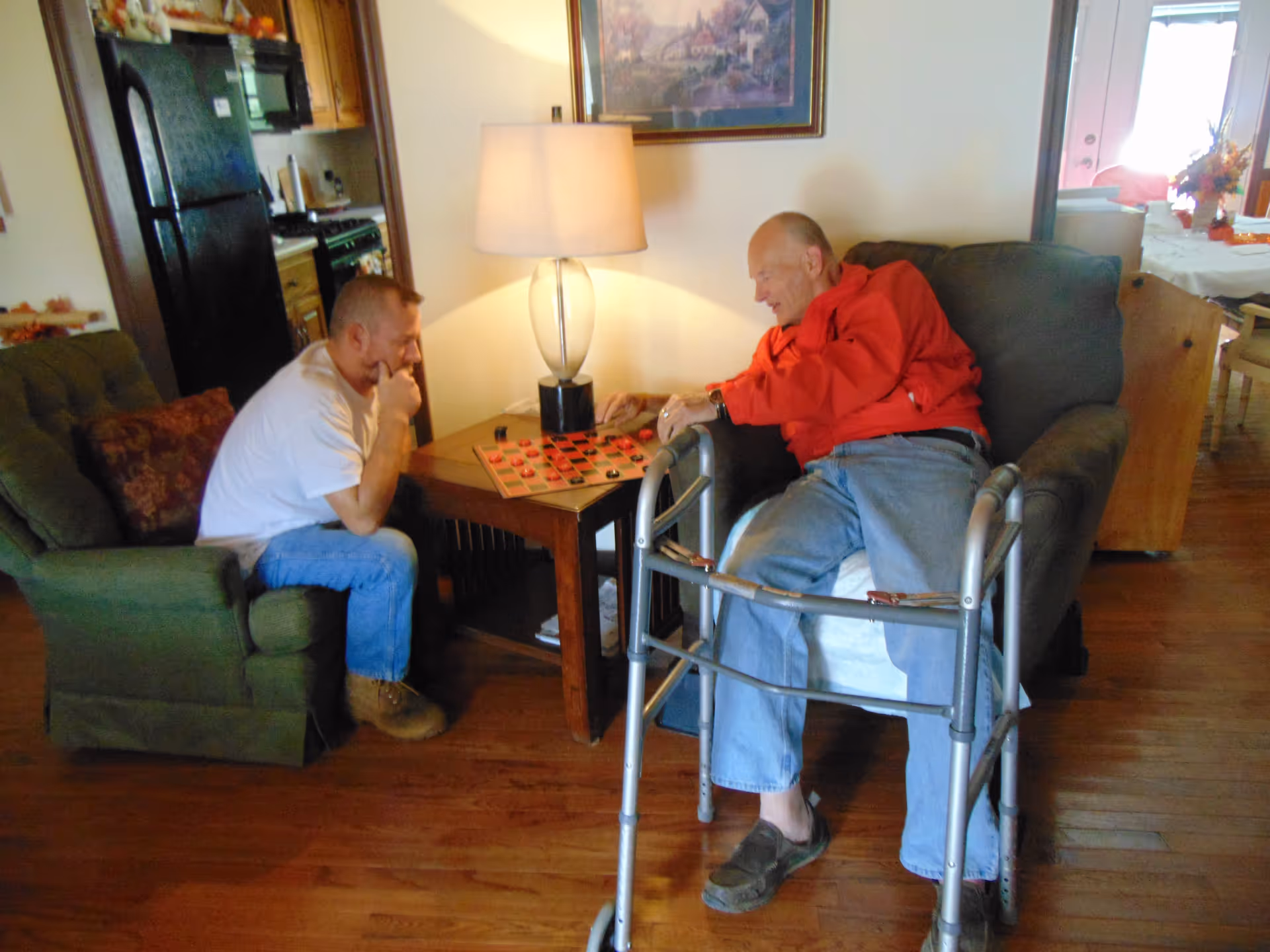 Two men playing checkers in a cozy living room area. One man is sitting in a green armchair, while the other man, wearing a red jacket and using a walker, is seated on a gray armchair. A wooden side table with a lamp and a framed painting on the wall are visible. The background shows a kitchen area and a dining table with decorations.