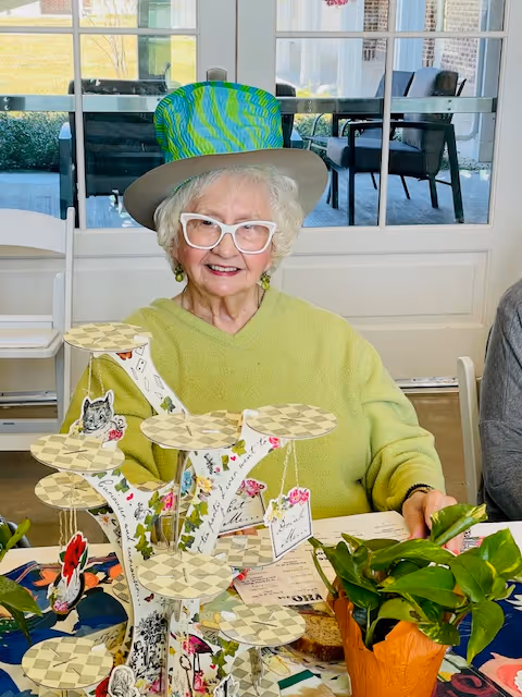 An elderly woman wearing a bright green sweater and a colorful tall hat sits at a table decorated with a multi-tiered display stand and a potted plant. She is smiling and wearing white-framed glasses. Behind her is a glass door leading to an outdoor patio with chairs and a table.