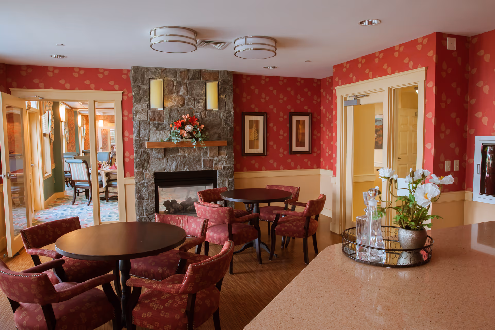 A cozy interior room with red patterned wallpaper and a stone fireplace decorated with a flower arrangement. There are two round wooden tables surrounded by red upholstered chairs. To the right, a countertop holds a decorative tray with a vase of white flowers and glassware. Through glass doors on the left, another room with dining tables and chairs is visible.