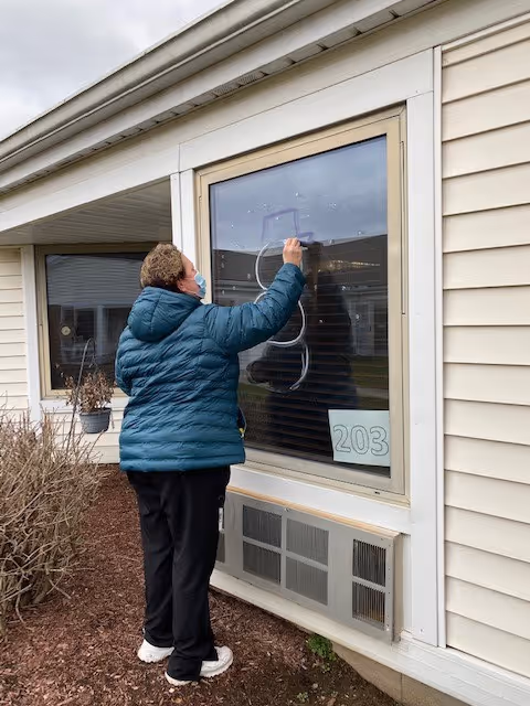 A person wearing a teal puffer jacket and black pants is writing or drawing on a window of a building. The window has the number 203 displayed on a paper inside. The building exterior is beige with siding, and there is an air conditioning unit below the window. Some bushes and mulch are visible near the building.