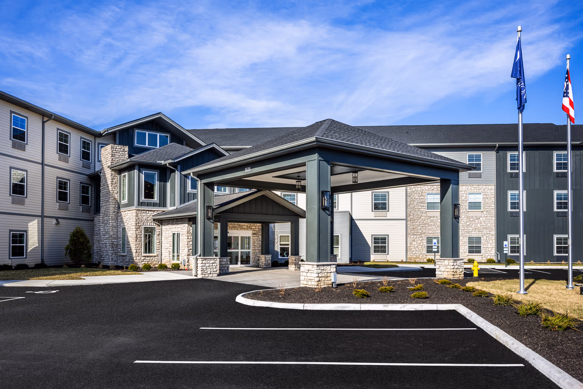 Exterior view of Provision Living at West Chester, showing a modern three-story building with a covered entrance, stone and siding facade, multiple windows, a parking area, and two flagpoles with flags against a blue sky.