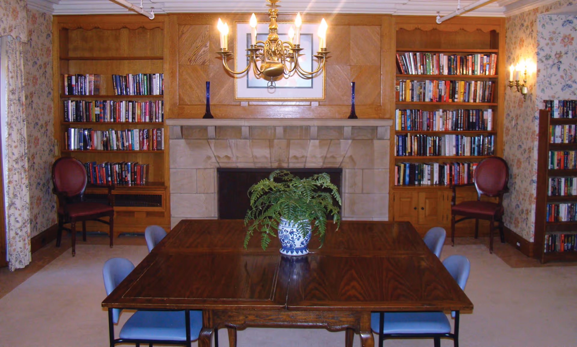 A cozy library or reading room with wooden bookshelves filled with books on either side of a stone fireplace. In front of the fireplace is a wooden table with a potted fern in a blue and white ceramic pot. Four blue chairs are placed around the table, and two red upholstered chairs are positioned near the bookshelves. The room has floral wallpaper, a chandelier hanging from the ceiling, and wall sconces providing light.