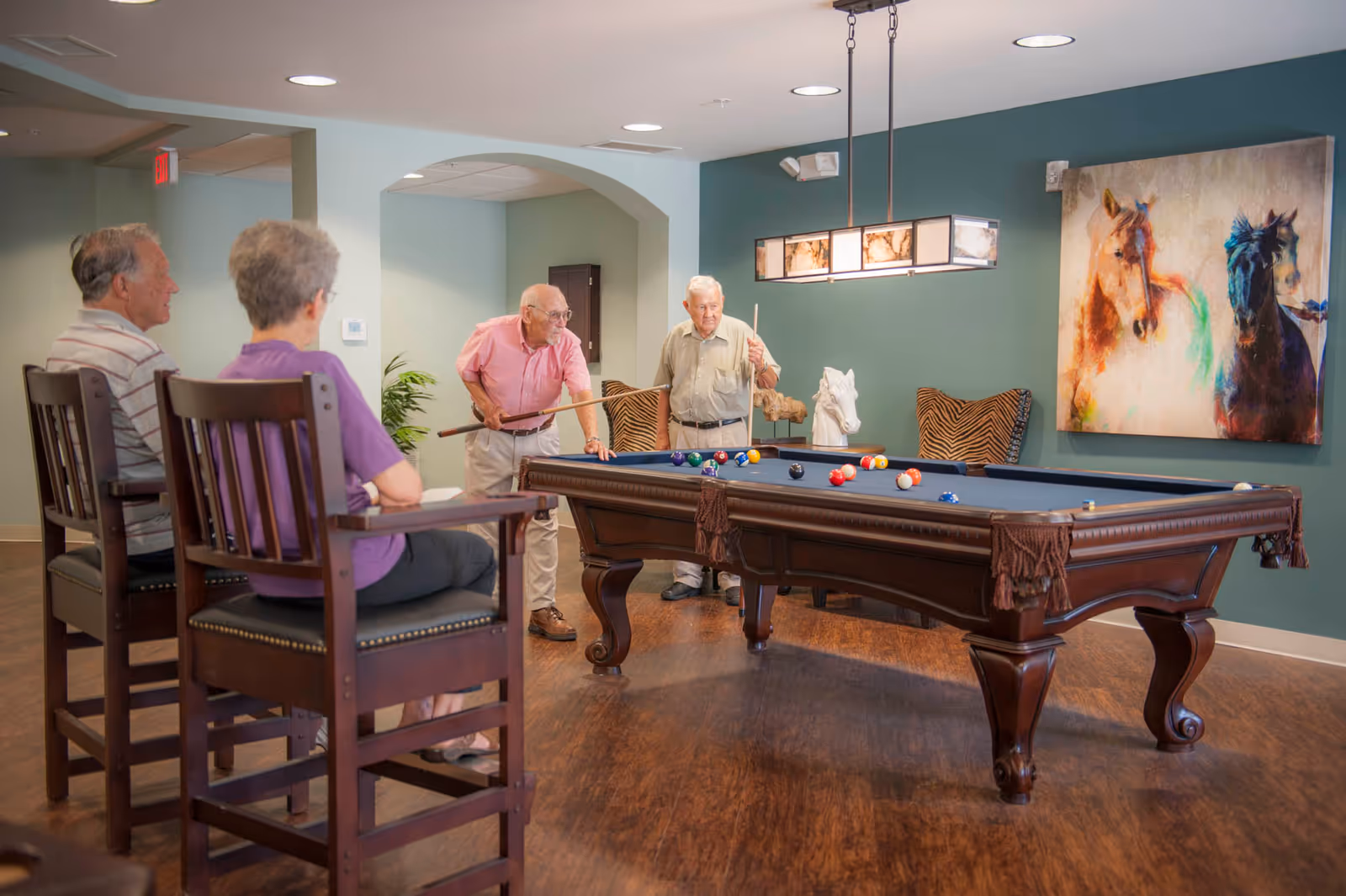 Two elderly men playing pool in a room with a blue pool table, while an elderly man and woman sit on wooden chairs watching. The room has wooden flooring, teal walls, a painting of two horses, and a modern rectangular ceiling light above the pool table.