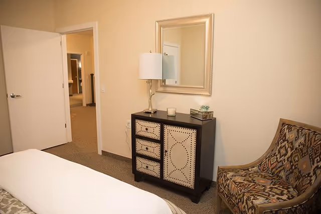 Cozy bedroom corner with a bed, patterned chair, a decorative dresser topped with a lamp and mirror, and an open door to a hallway.