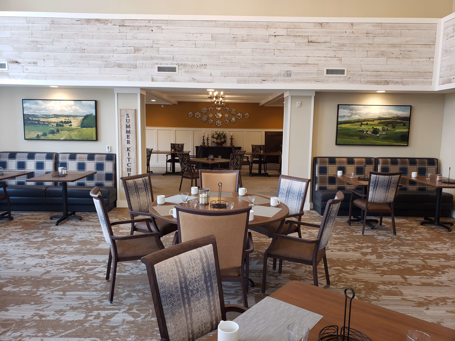 A senior living facility dining room with round and rectangular wooden tables surrounded by cushioned chairs. The walls are decorated with landscape paintings and a sign that reads 'SUMMER KITCHEN'. The room has a warm, inviting atmosphere with carpeted floors and soft lighting.