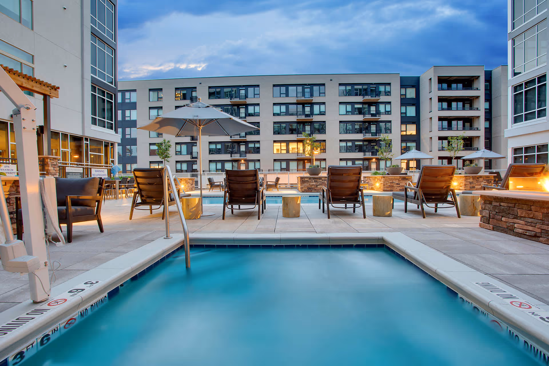 Outdoor pool area at a senior living facility with lounge chairs, umbrellas, and a modern multi-story apartment building in the background during early evening.