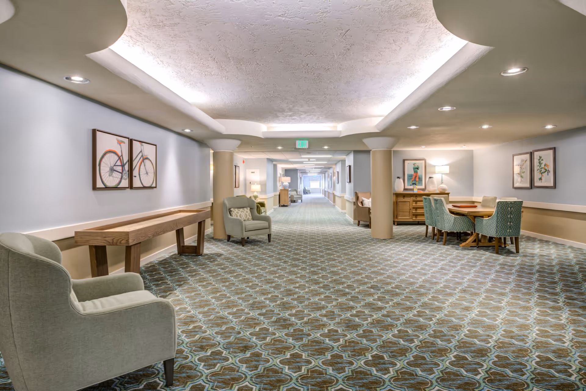 A long, carpeted hallway in a senior living facility with light blue walls and beige accents. The hallway features several armchairs, a wooden table, framed artwork on the walls, and a small dining table with four chairs. The ceiling has recessed lighting and a textured design.