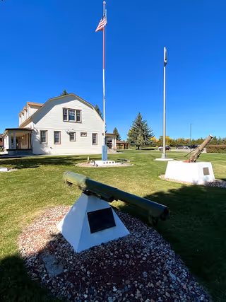 Outdoor view of a grassy area at Veterans' Home of Wyoming featuring two cannons mounted on white concrete bases, two flagpoles with one flying the American flag, and a large white building with multiple windows under a clear blue sky.