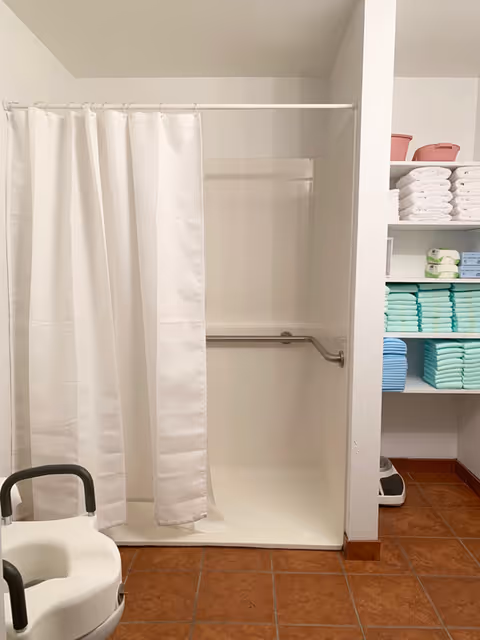 Accessible shower stall with a white curtain and grab bar, a shower chair in front, and shelves of folded towels and linens nearby.