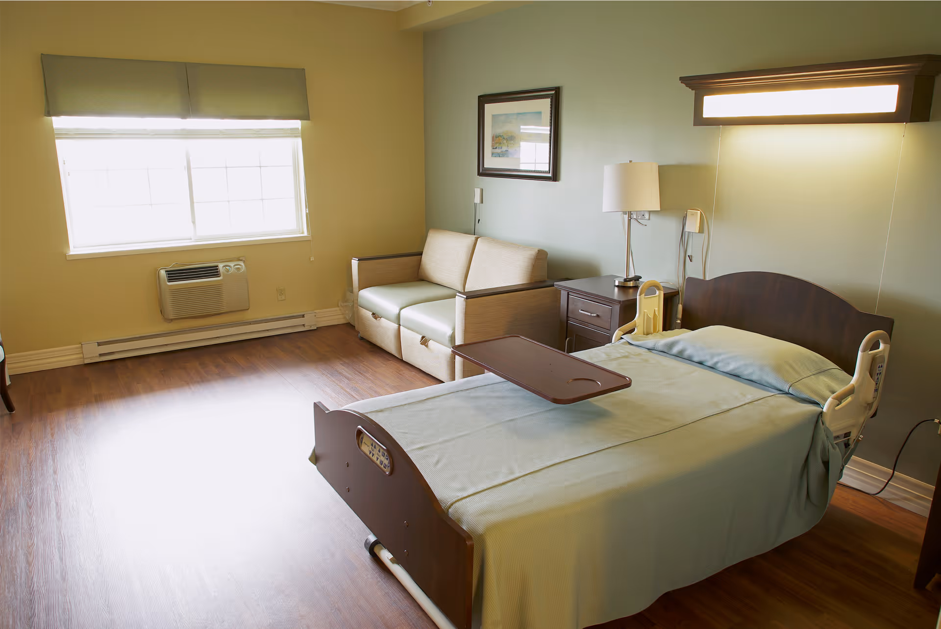 A well-lit room with a hospital-style bed covered in a light green blanket, a wooden bedside table with a lamp, a small beige sofa, and a large window with blinds and a green valance. The floor is wooden, and there is a wall-mounted air conditioning unit below the window.