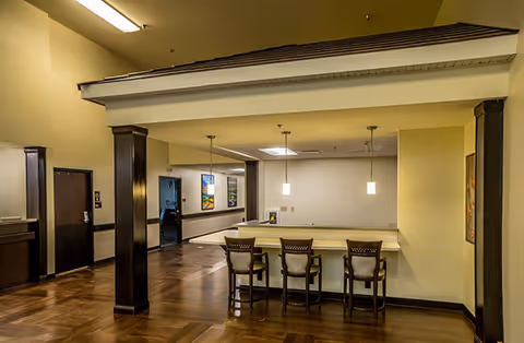 Interior view of a common area in a senior living facility featuring a counter with three high chairs, pendant lights hanging above, dark wood flooring, and beige walls with dark wood trim. There are doorways and framed artwork visible in the background.