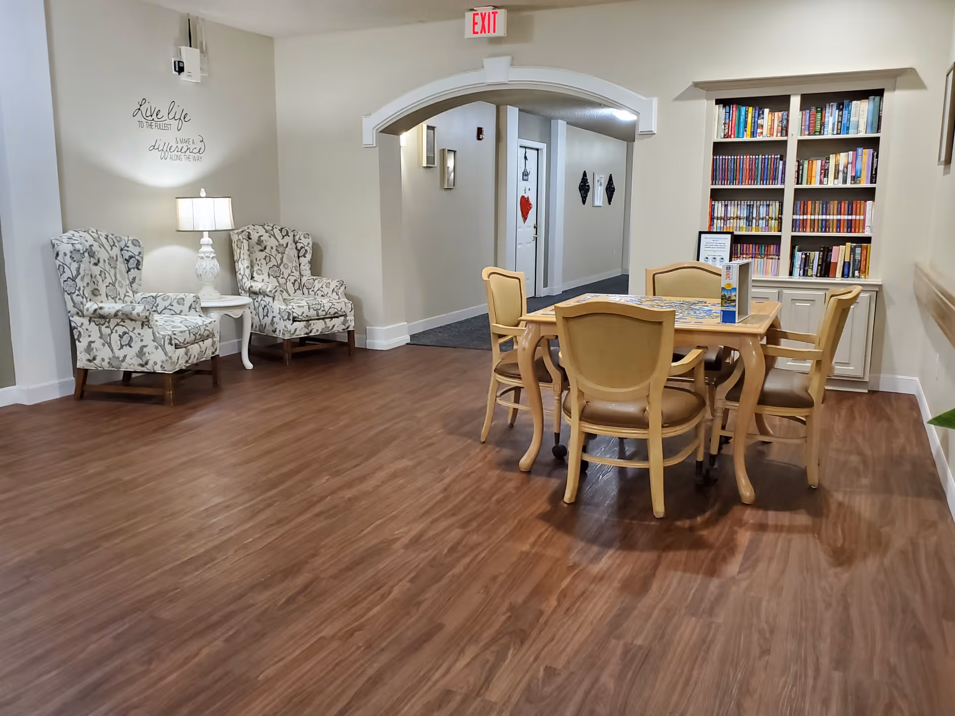 A cozy interior common area with two floral armchairs and a small white side table with a lamp on the left. In the center, there is a wooden table with four beige chairs around it, and a puzzle on the table. On the right side, there is a built-in bookshelf filled with books. The floor is wood, and there is an archway leading to a hallway in the background. A wall decal reads 'Live life to the fullest & make a difference along the way.'