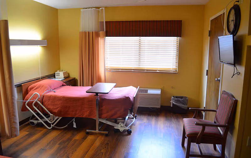 Sunlit patient room with a hospital-style bed covered in a red blanket, adjustable overbed table, armchair, wall-mounted TV, and a window with blinds.