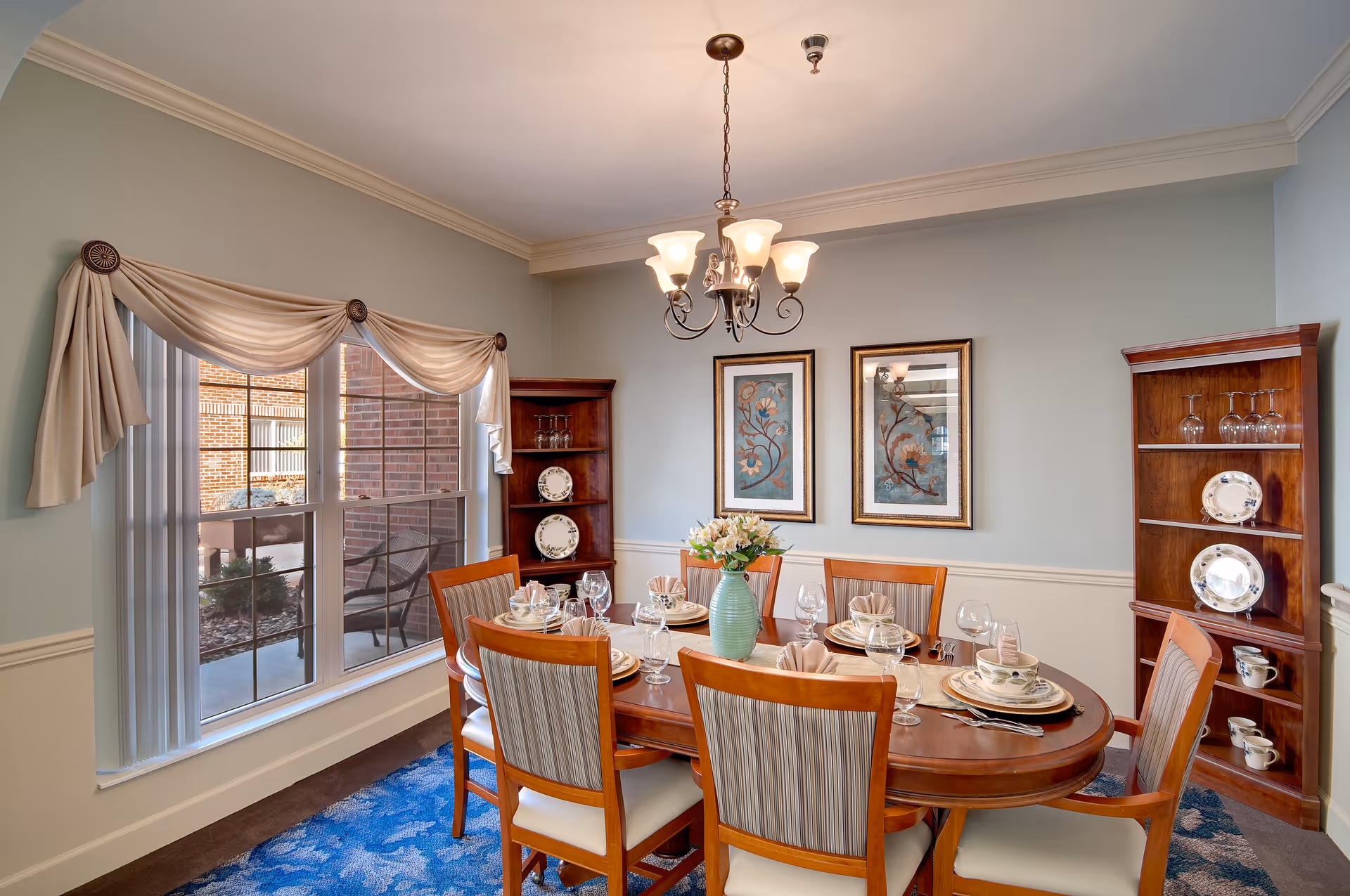 A well-lit dining room with a wooden dining table set for six people. The table is arranged with plates, bowls, glasses, and napkins, and a green vase with flowers serves as a centerpiece. The room features two wooden corner shelves displaying plates and glassware, light blue walls with white wainscoting, two framed floral artworks, a window with cream-colored drapes, and a chandelier hanging from the ceiling.