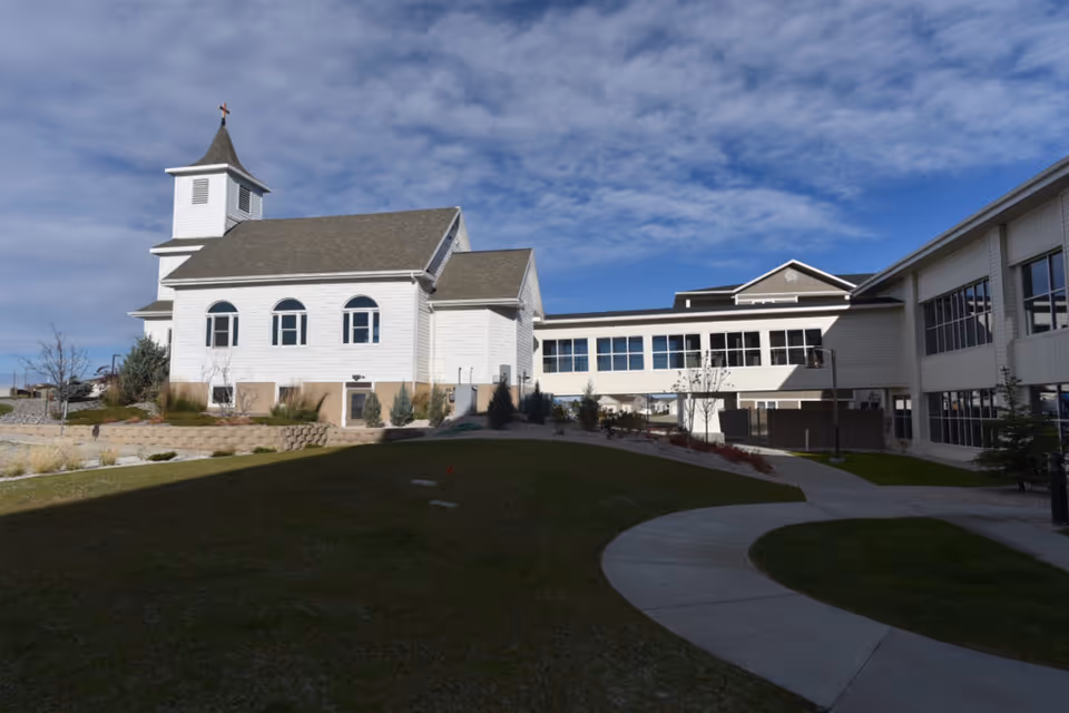 White chapel-like building connected to a senior living facility with a curved sidewalk and lawn under a partly cloudy sky.