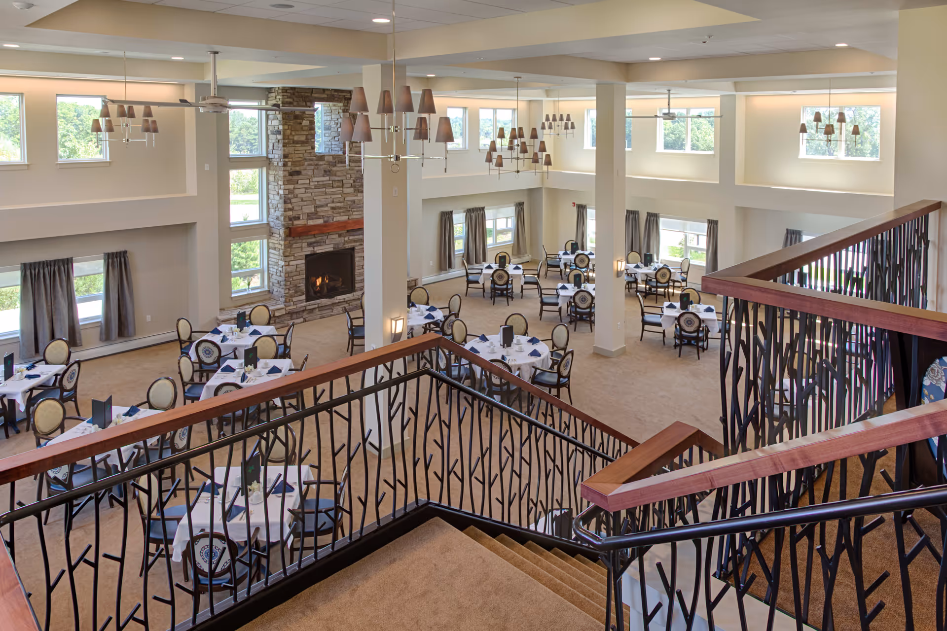 Spacious dining room with multiple round tables set with white tablecloths, blue napkins, and chairs with patterned backs. The room features large windows, a stone fireplace, beige carpet, and modern chandeliers hanging from the ceiling. A staircase with decorative black railings and wooden handrails is visible in the foreground.